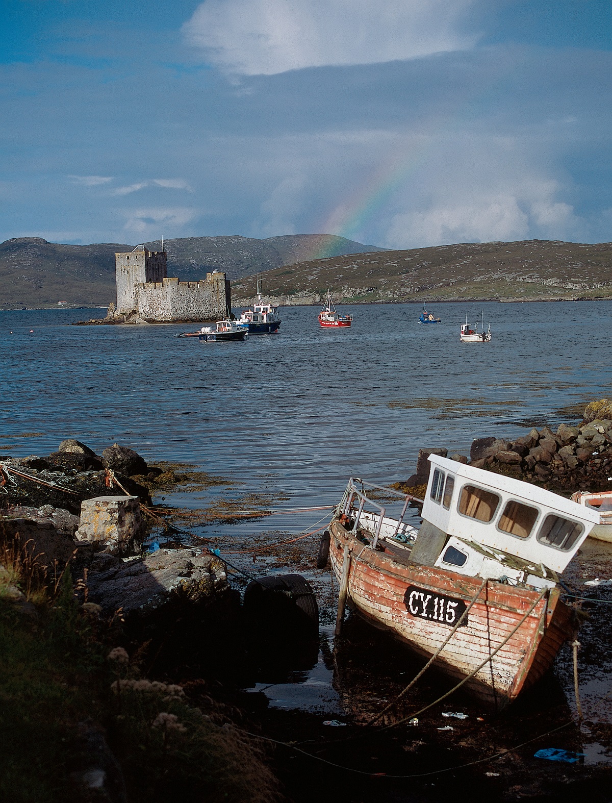 A wide bay with a stone castle on a small island in the distance, colourful boats on the water, and an old wooden fishing boat on the rocky shore in the foreground. A faint rainbow arcs above the hills.