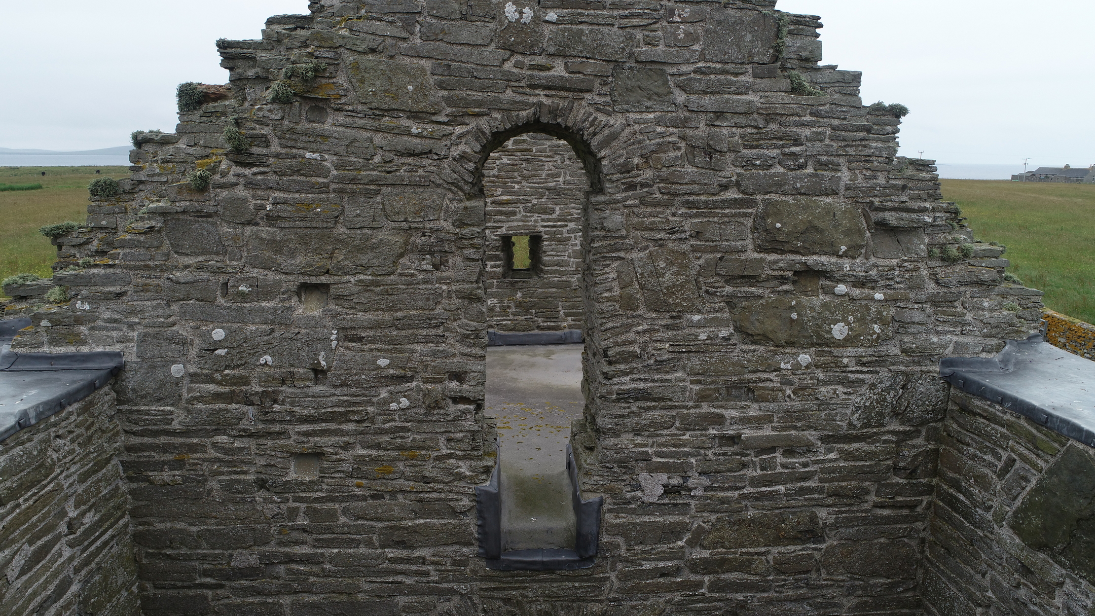 Stone gable wall of the church with arched windows and narrow openings, viewed from inside the ruin.