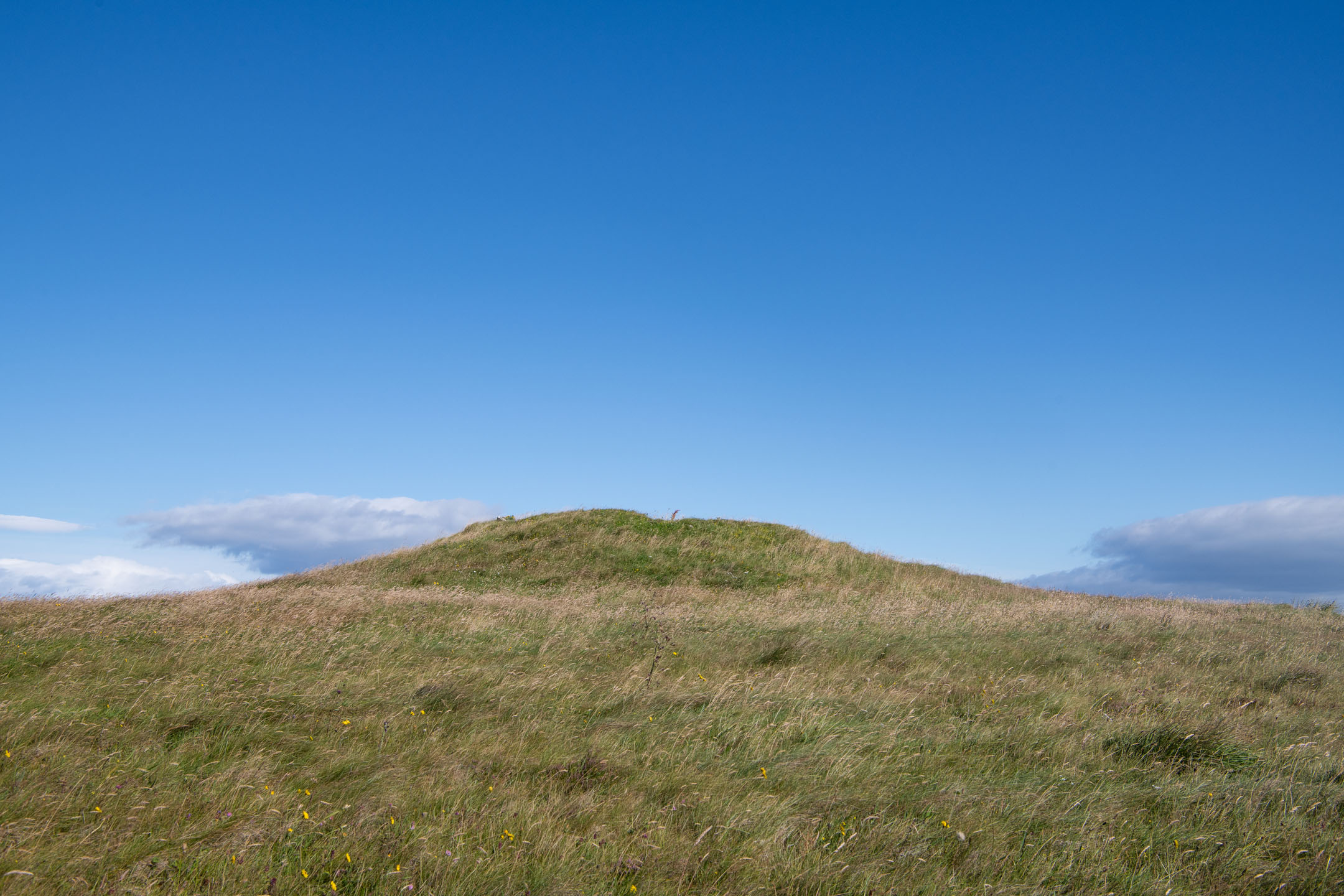 Low grassy cairn with blue sky and distant clouds.