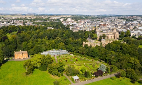 Aerial view of Dunfermline showing extensive green parkland, formal gardens, and tree‑lined grounds. A large historic stone abbey complex sits to the right, with a glass‑roofed greenhouse and an orange‑coloured manor-style building visible to the left. The town’s buildings stretch out across the background under a bright, partly cloudy sky.