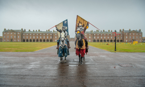 Two knights in armour sits atop horses holding up heraldry flags. Behind them is a grass field and a large brick building in the background. A jousting arena can be seen on the right.