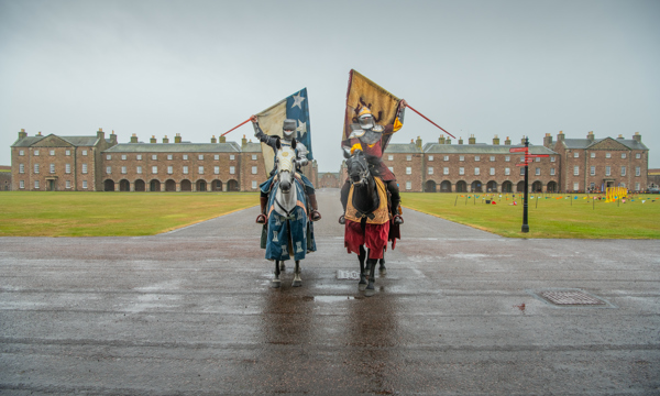 Two knights in armour sits atop horses holding up heraldry flags. Behind them is a grass field and a large brick building in the background. A jousting arena can be seen on the right.