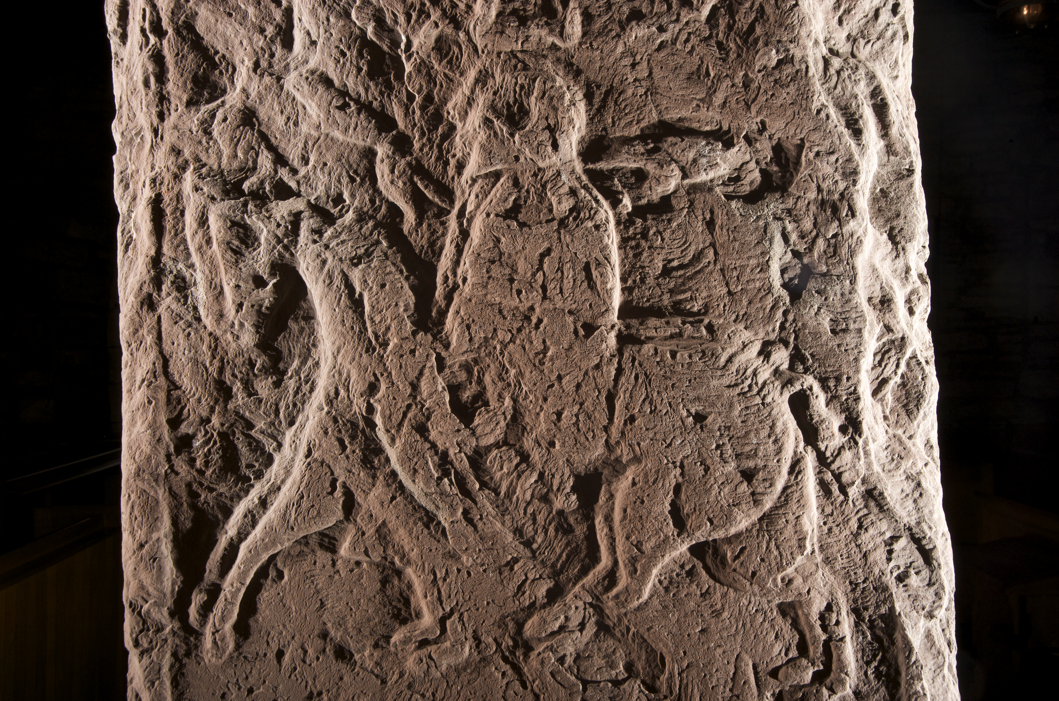 Detailed view of a sculptured stone featuring a central figure on horseback, surrounded by other carved elements including animals and abstract patterns. The carvings are deeply incised into the stone, which has an uneven, aged surface. Lighting emphasizes the contours and shadows of the artwork.