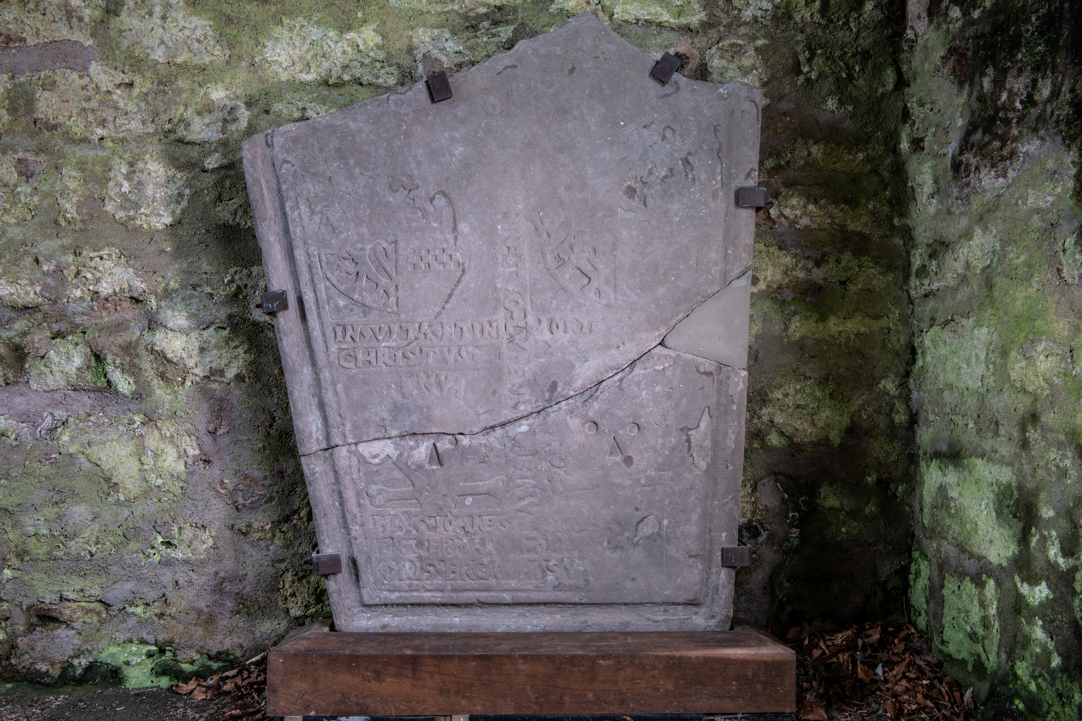 Weathered stone grave slab mounted on a wooden base and secured with metal brackets, featuring carved heraldic shields and partially legible inscriptions. The slab is cracked horizontally and vertically, with moss-covered stone walls in the background.