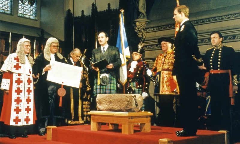 A photograph of a stone on a table, surrounded by people reading from a book.