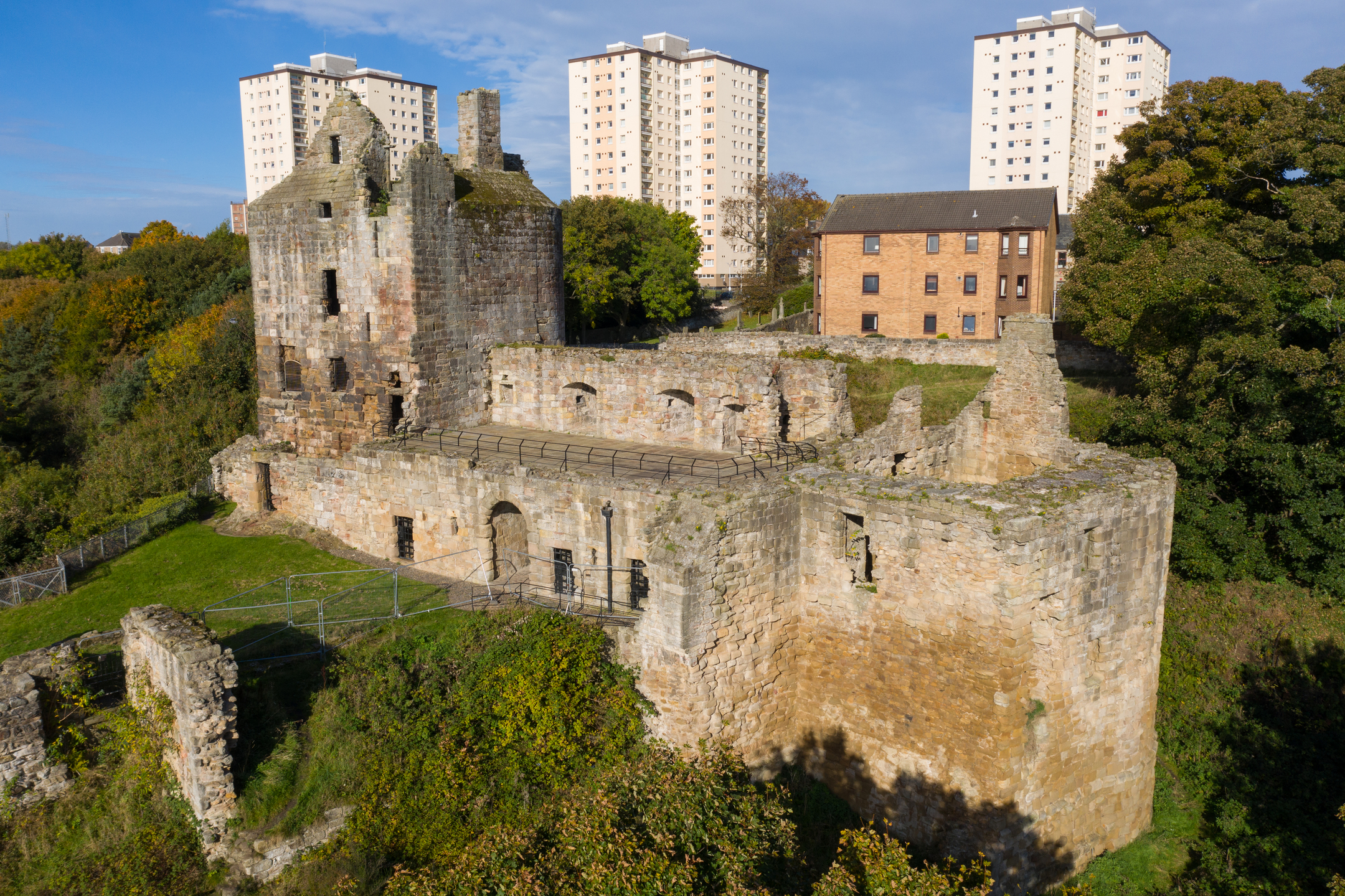 An aerial view of Ravenscraig Castle’s long stone façade and collapsed outer walls, with several tall modern tower blocks rising in the distance. The medieval ruins sit among green trees and grassy banks under a bright sky, highlighting the contrast between historic and contemporary architecture.