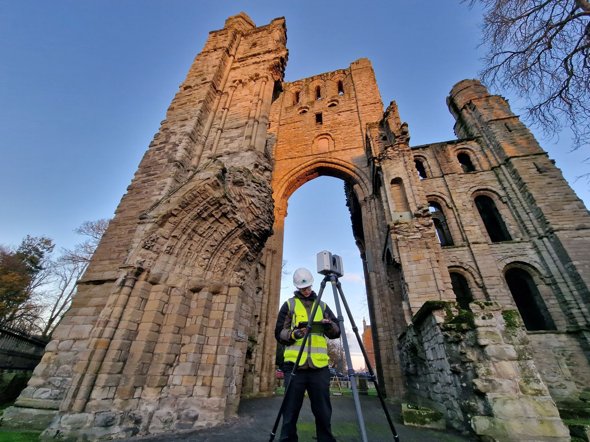 Survey equipment set up in front of towering west front ruins under clear sky.