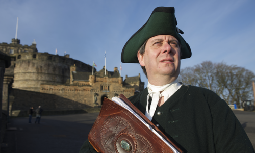 A man in a tricorn hat, white shirt and black cloak stands facing the camera and looking off to the right. He holds a large leatherbound book. Edinburgh Castle, a large stone castle with four flags, is in the background.