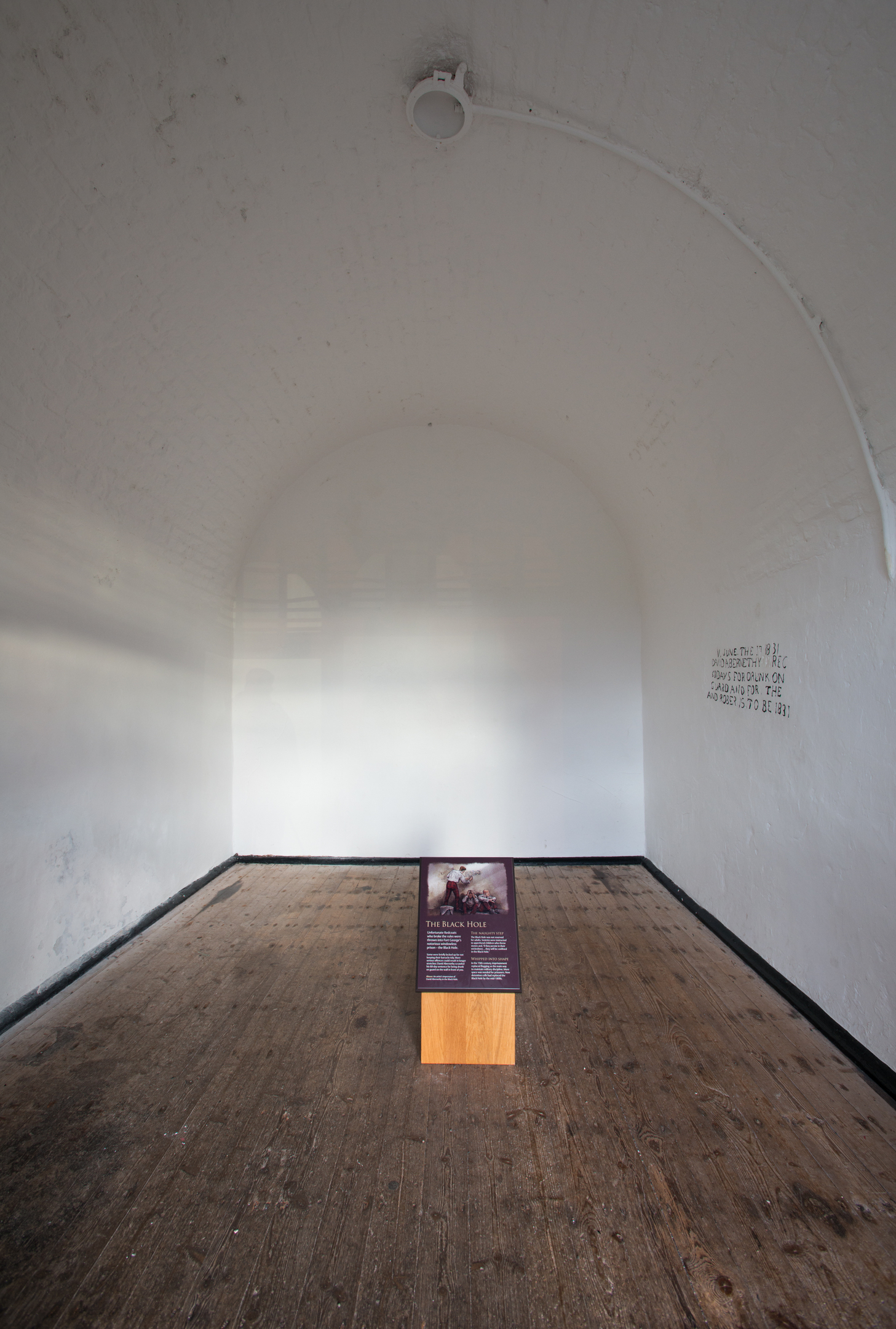 A small white‑walled vaulted room at Fort George containing a single display panel on a wooden plinth in the centre.