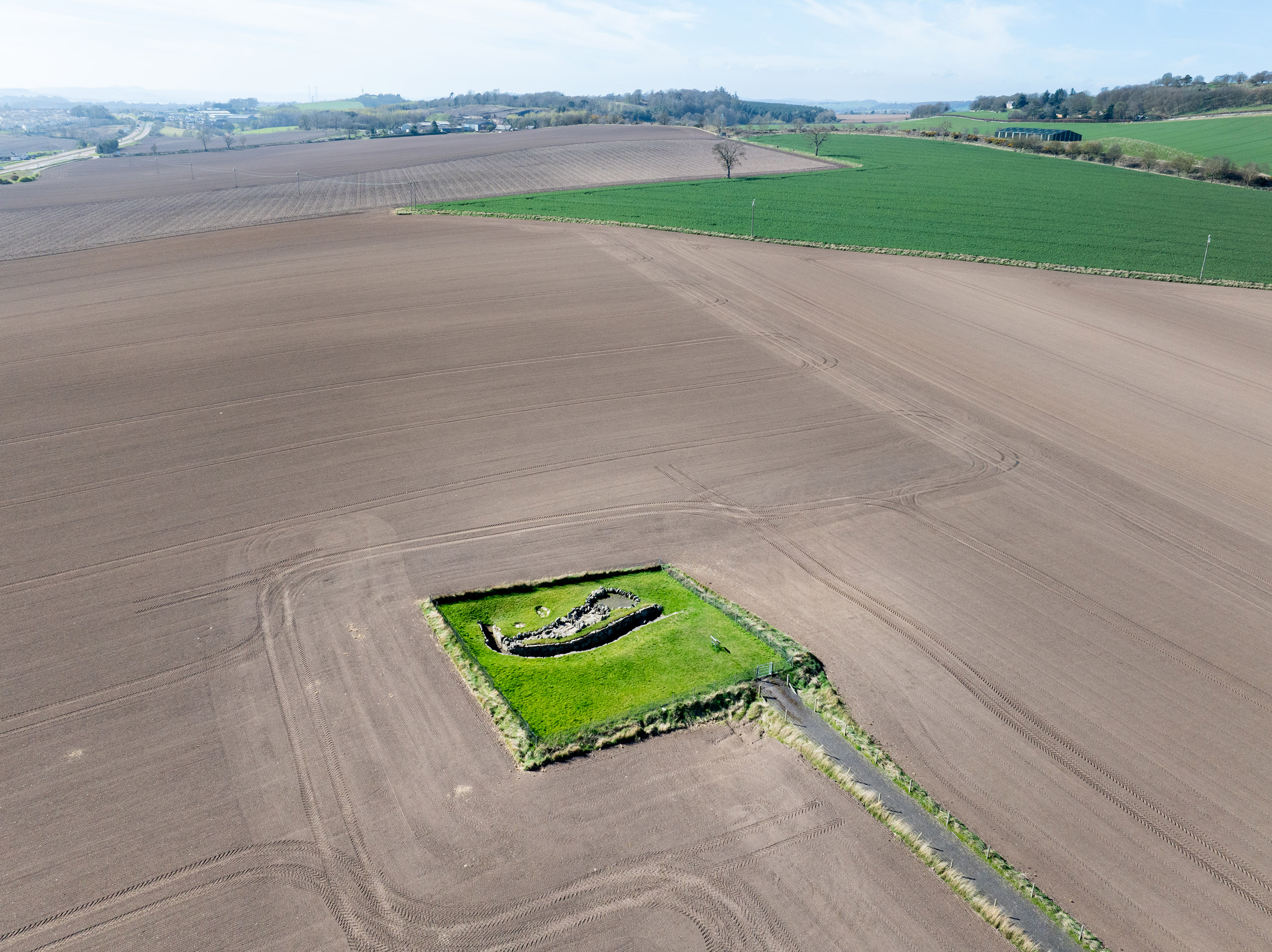 A distant aerial view of Ardestie Earth House, showing the small, rectangular grass enclosure containing the stone remains of the ancient underground structure, surrounded by wide expanses of ploughed brown farmland. Green fields and gentle hills stretch across the background under a lightly clouded sky.