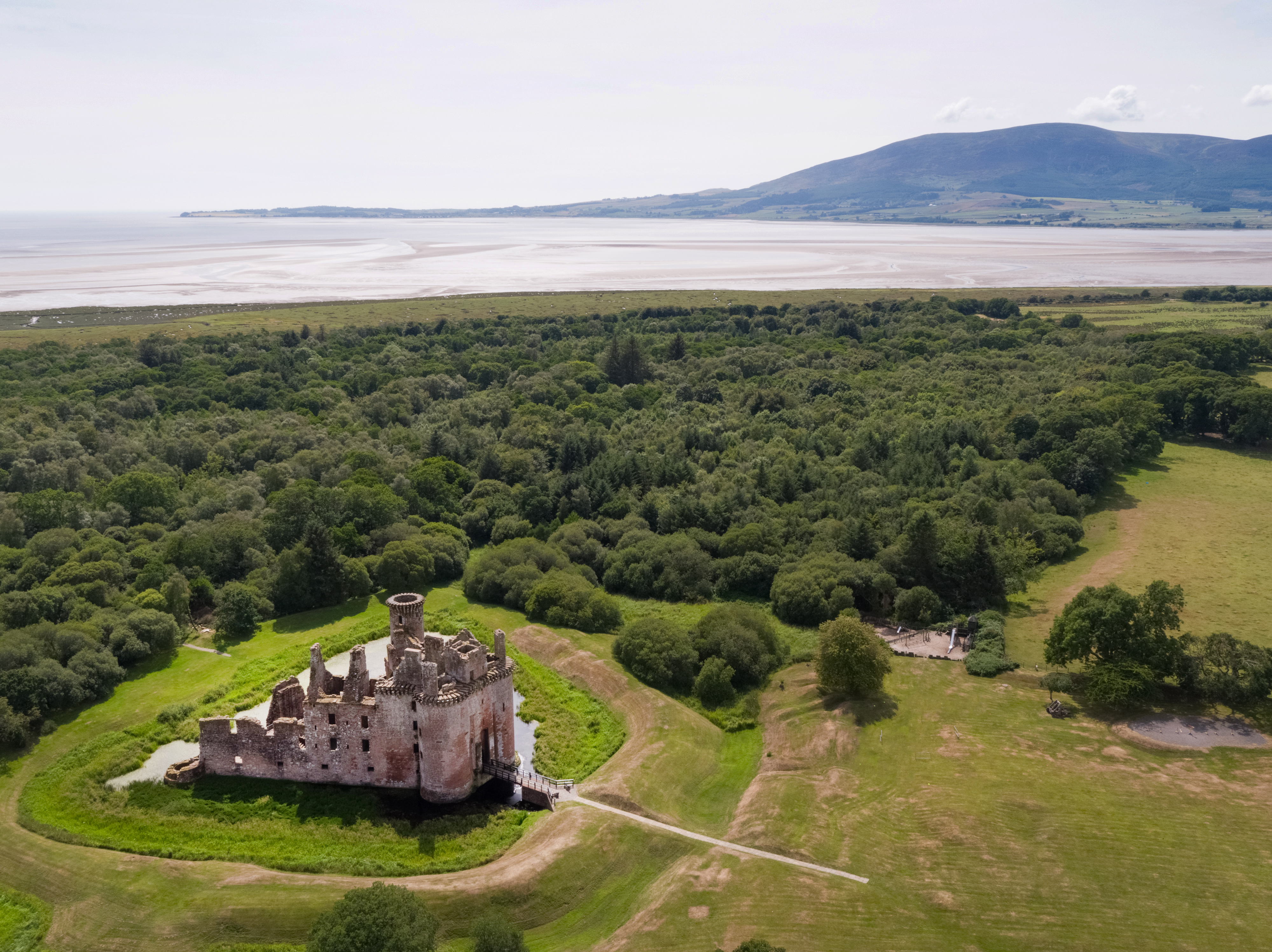 An aerial view of a large ruined stone castle surrounded by a moat and set within open grassland. Dense green woodland stretches out behind the castle, leading to a wide sandy shoreline and calm water in the distance. Low hills rise on the far side of the bay
