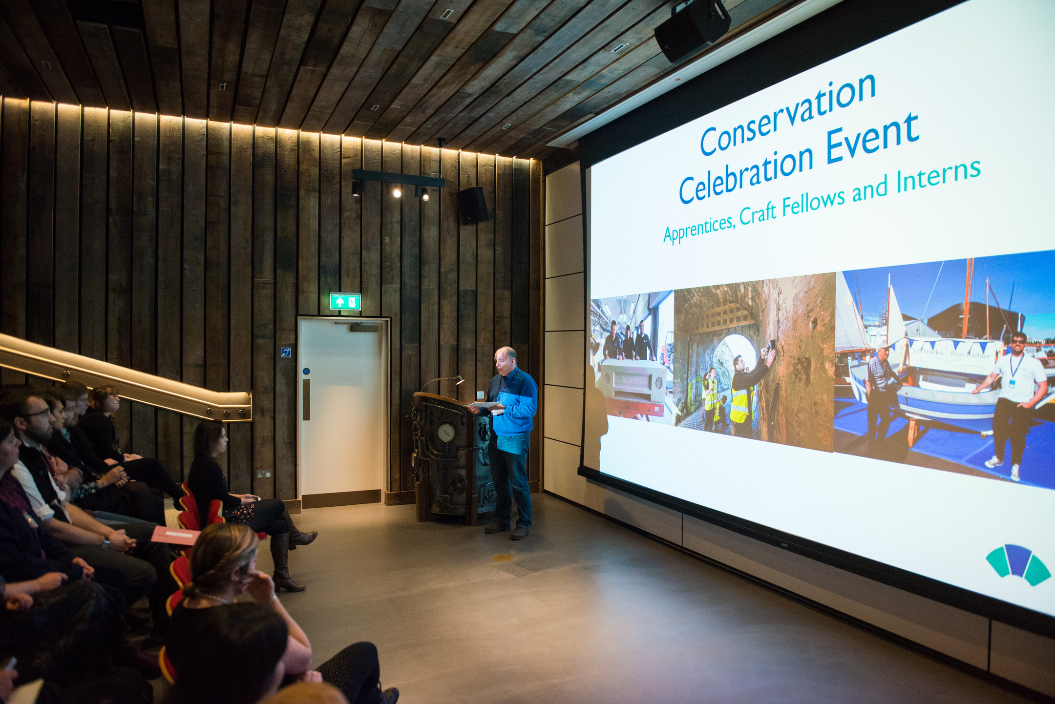 An audience watches a presentation with an opening slide that says 'Conservation celebration event'