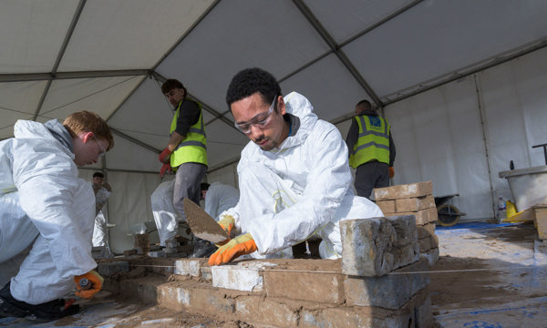 Students in white overalls, wearing safety glasses and gloves while learning to build a wall
