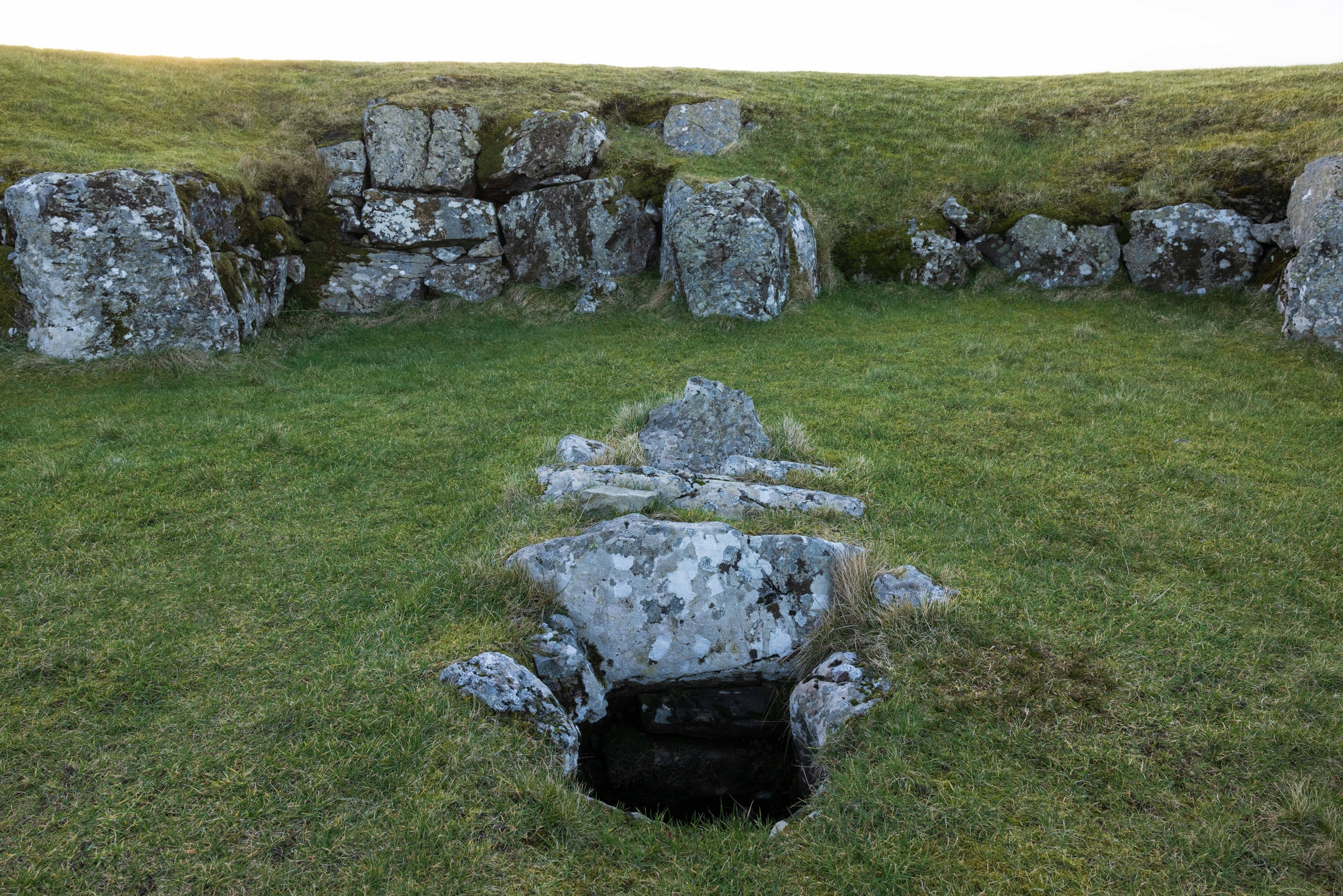 Close-up of a stone-lined pit within the grassy interior of Stanydale Temple. The pit is rectangular and partially covered by flat stones, located near the center of the oval enclosure. Large upright stones form the surrounding wall.
