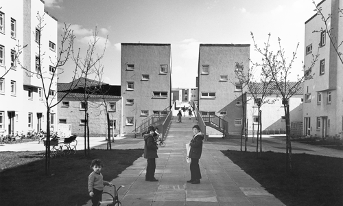 A black and white photograph showing two people stood on a paved courtyard. There are patches of grass with bare trees on either side of them and a child with a tricycle in the foreground. Large square buildings with lots of windows surround the courtyard.