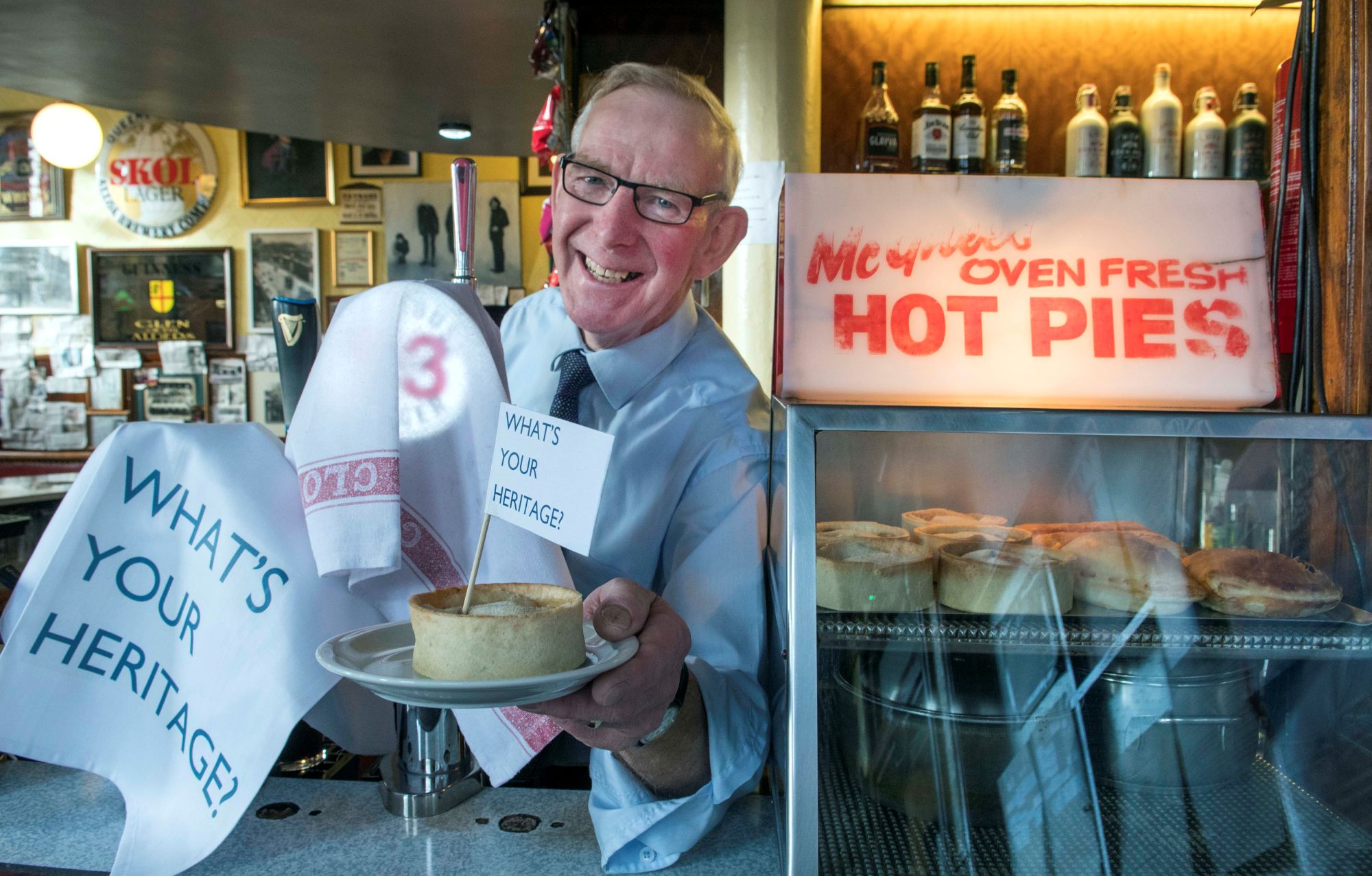 Older man, wearing glasses and a blue button up shirt and tie, stands behind a bar holding out a pie with a flag in it that reads 'What's your heritage'.