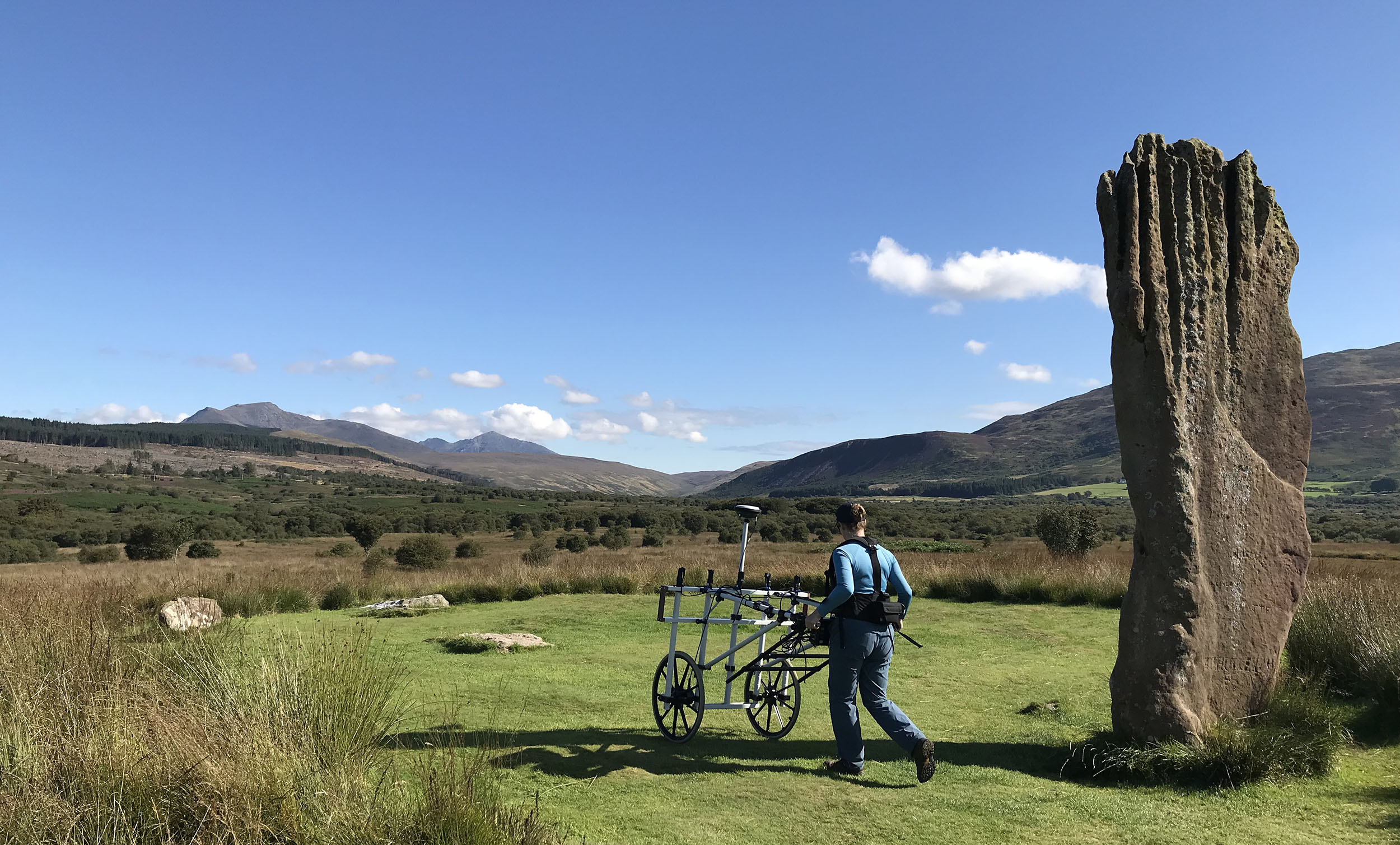 A person conducting a geophysical survey with a wheelbarrow-like instrument near a standing stone