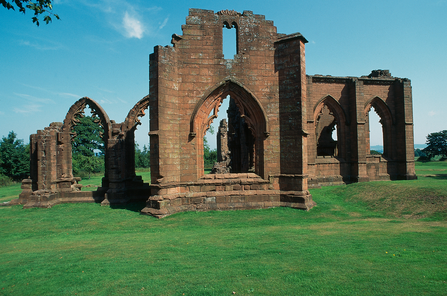 Remains of stone historical ruins, with multiple arches, with detailing around the edges of it. It sits on a grass landscape. 