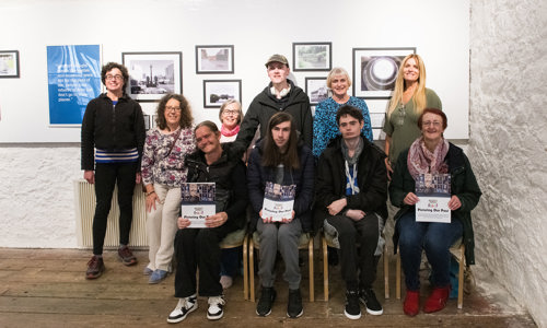 A group of ten people, four sitting in chairs and five standing behind them, pose in front of a white wall which has ten framed black and white photographs of various sizes hanging on it. They look to the camera smiling, with some holding a booklet labelled Picturing Our Past.