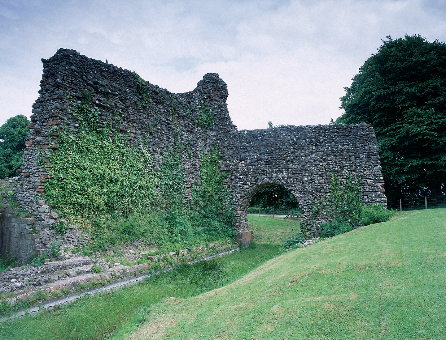 Two connecting large stone walls, which looks like a corner of the remains of an old castle. There is an arch, with a path running through the centre. There is a tree in the background. 