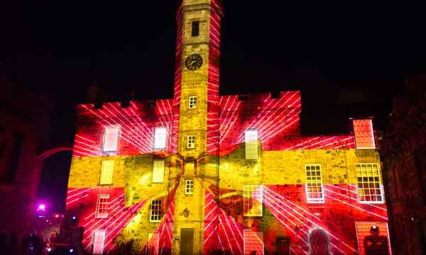A photo of a stone clock tower in Crown Square in Edinburgh Castle at night, with colourful projections depicting red wrapping paper and a large yellow ribbon making the building appear as though it's a wrapped gift.