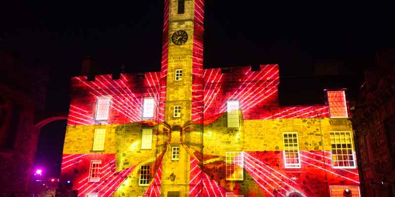 A photo of a stone clock tower in Crown Square in Edinburgh Castle at night, with colourful projections depicting red wrapping paper and a large yellow ribbon making the building appear as though it