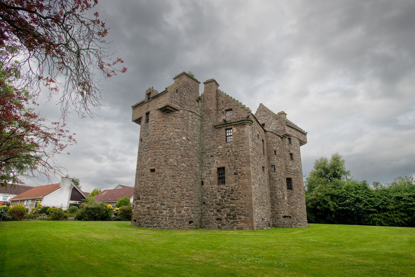 A stone castle, with multiple small windows on it. There is grass in front of castle, the sky is grey.