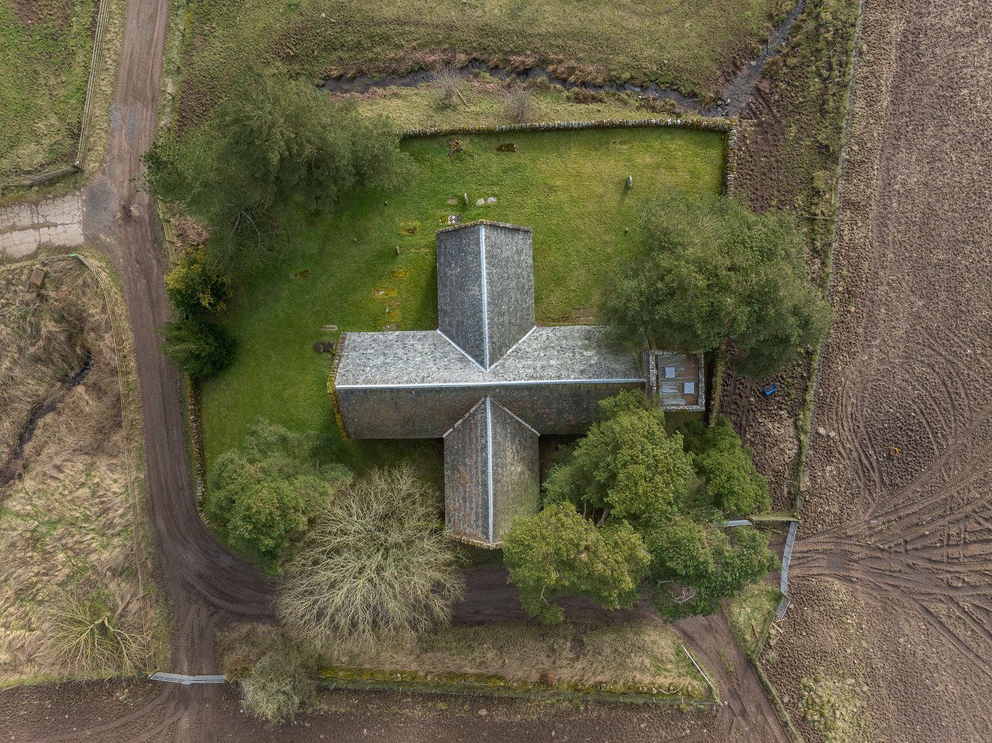 aerial view looking directly down onto a small stone church shaped like a cross, set within a walled grassy enclosure and surrounded by trees. A narrow track runs along two sides of the site, with ploughed fields and a small stream bordering the remaining edges.
