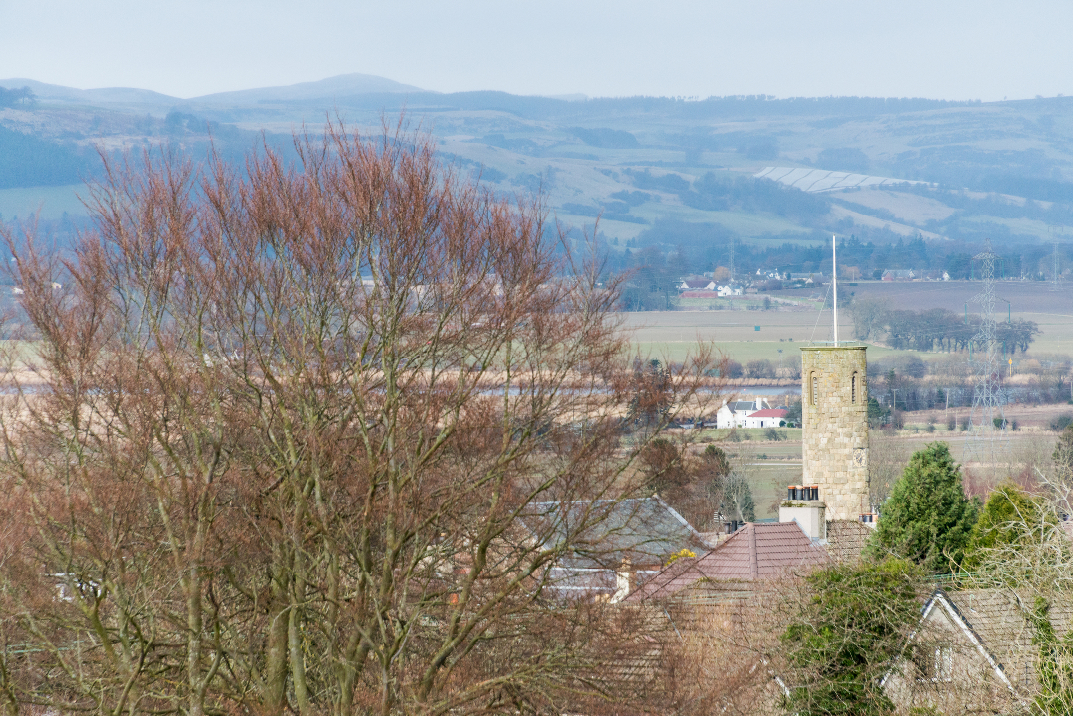 A distant view of Abernethy Round Tower rising above village rooftops, with bare trees and layered hills stretching into the background.