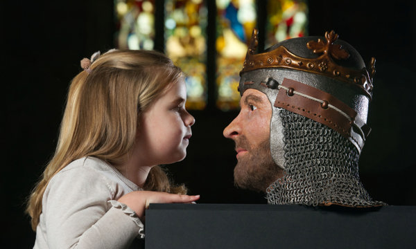 A girl in Dunfermline Abbey looking at a 3D model of Robert the Bruce's face, with a stain glass window in the background