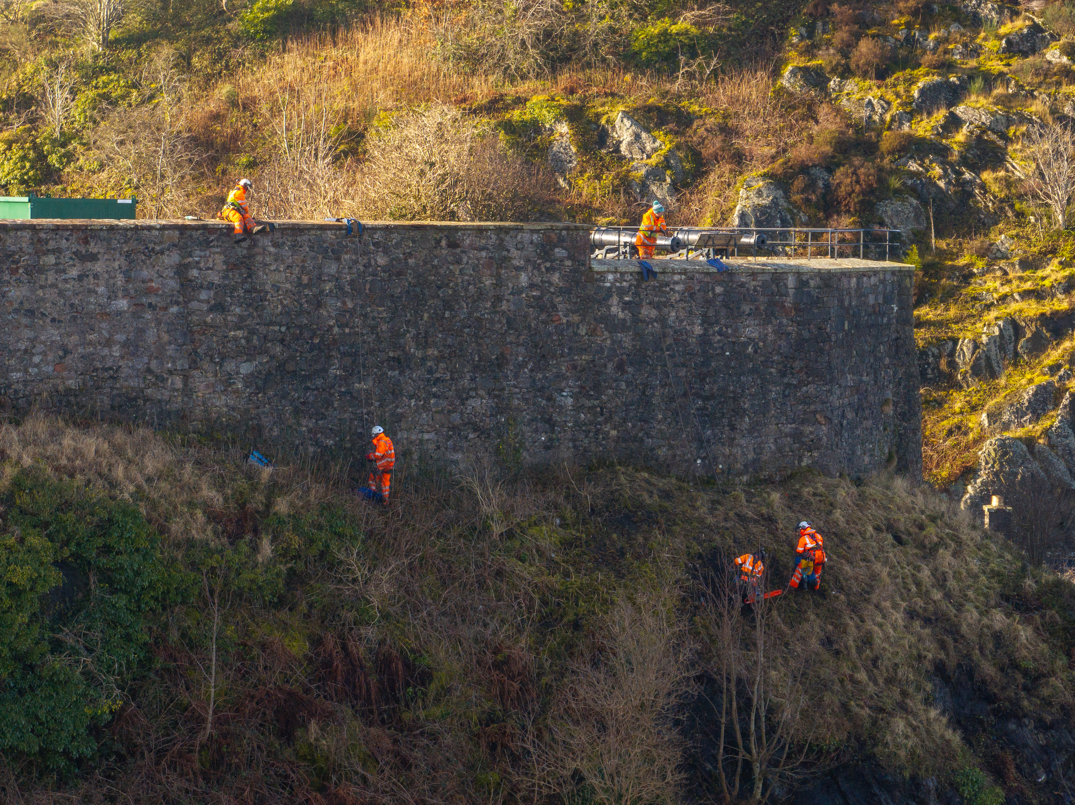 A group of workers in orange hi-vis jackets inspecting masonry on a castle wall. To do this, they have had to abseil over the battlements.  