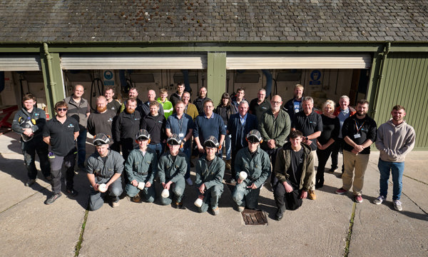 Group of around 30 people stand in the sunshine in front of workshop. A number of the group are in uniform and protective clothing. Those crouching in the front row hold stonemasonry tools.