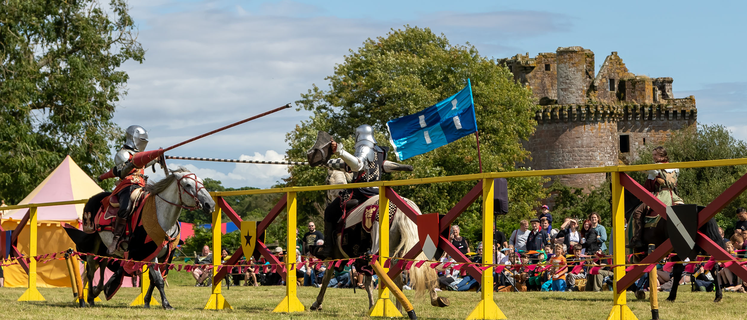 Two knights on horseback charge at each other with lances in hand, in front of a crowd of people. Caerlaverock Castle sits in the background