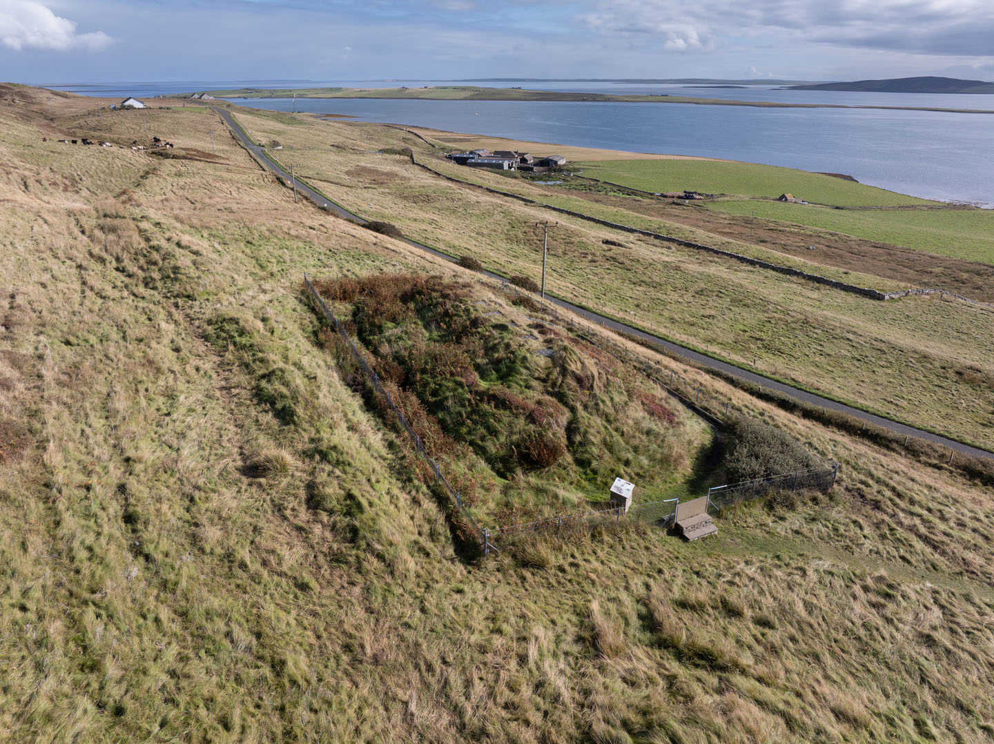 Aerial side view of the grass-covered, mounded cairn with fenced entrance and shore landscape in background.