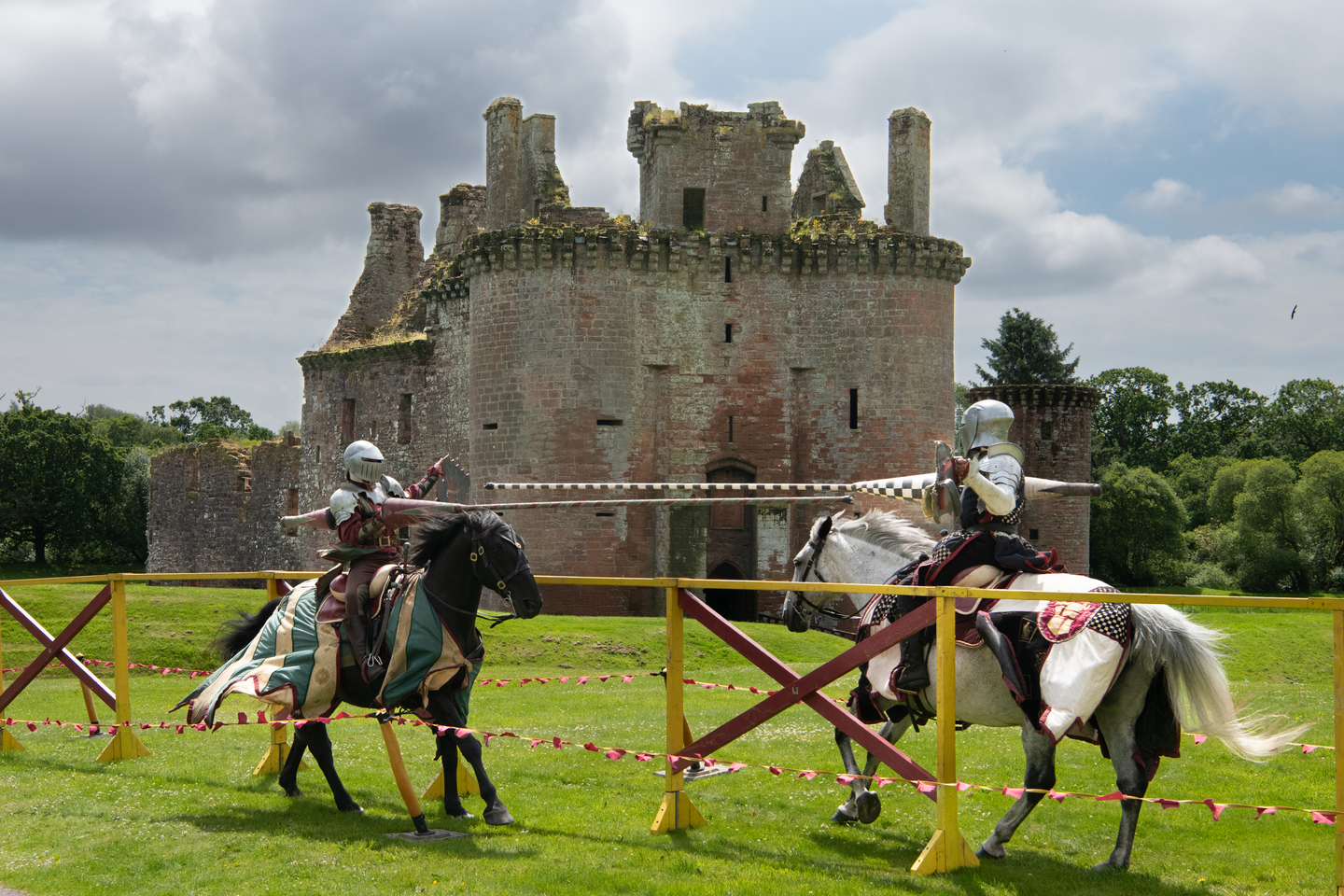 two knights on horseback face one another with lances across a yellow barrier, in front of a stone castle 