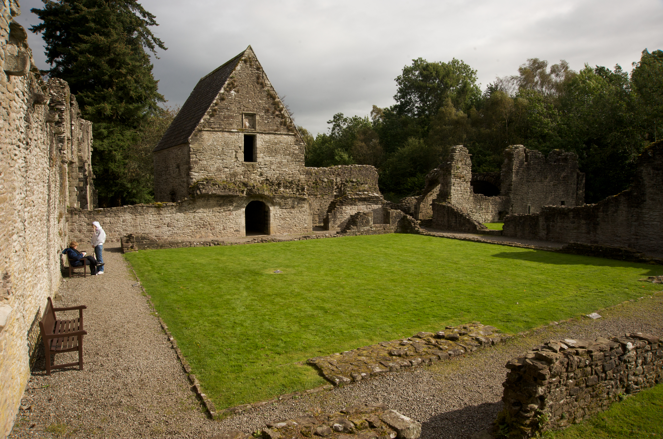 A grass courtyard enclosed by stone walls and ruined buildings. It is flanked by a gravel path and wooden benches. 