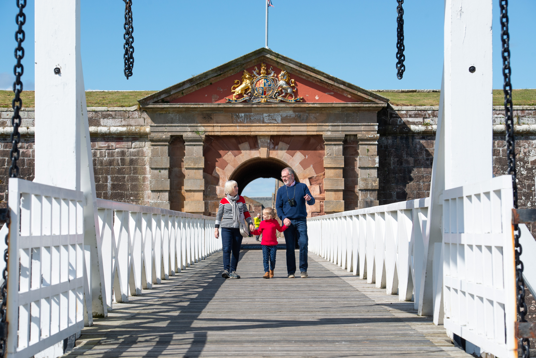 A family walking hand‑in‑hand across the white wooden bridge leading to Fort George’s main gatehouse, decorated with a royal crest.