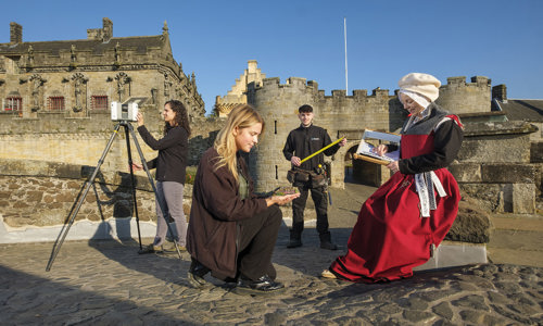 Four people are arranged in various poses in front of Stirling Castle, it is a clear blue sky day. In the centre, a blonde girl crouches down on one knee, facing right holding a miniature building in her hand. In front of her another women is sitting dressed in period clothing. In the middle ground between them and the castle, a young man hold open a measuring tape, he has a tool belt hanging from his waist. And the final person is on the left, they are looking into an instrument that is on a tall tripod.