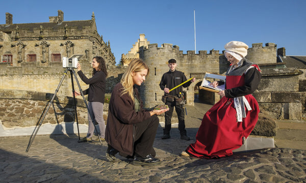 Four people are arranged in various poses in front of Stirling Castle, it is a clear blue sky day. In the centre, a blonde girl crouches down on one knee, facing right holding a miniature building in her hand. In front of her another women is sitting dressed in period clothing. In the middle ground between them and the castle, a young man hold open a measuring tape, he has a tool belt hanging from his waist. And the final person is on the left, they are looking into an instrument that is on a tall tripod.