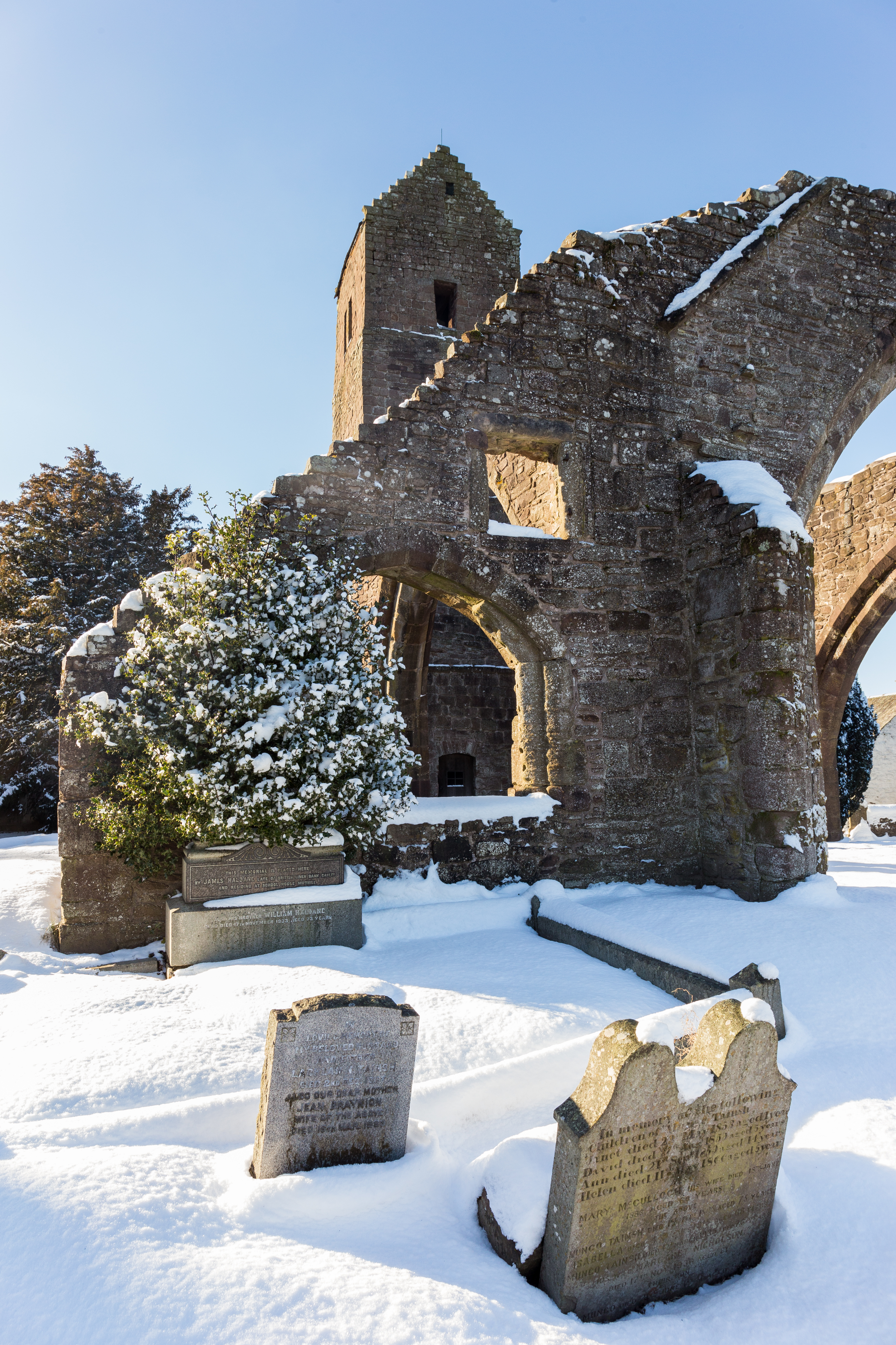 Snow-covered gravestones in front of the arched ruins of Muthill Old Church, with bright sunlight shining through the stone arches.
