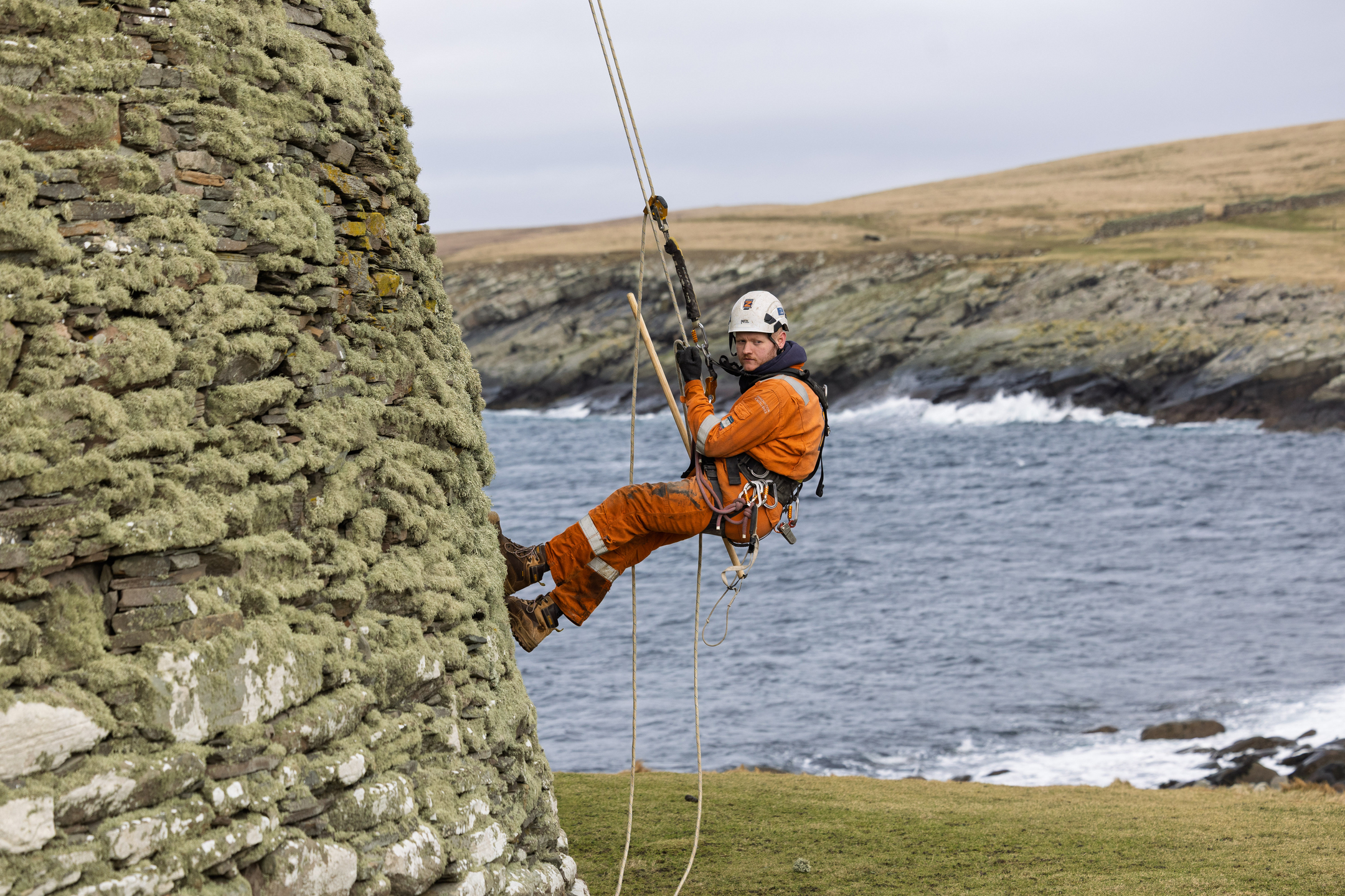 Rope‑access conservator in orange protective clothing and a safety helmet suspended on ropes beside the stone wall of Mousa Broch, with the rocky shoreline and sea visible in the background.