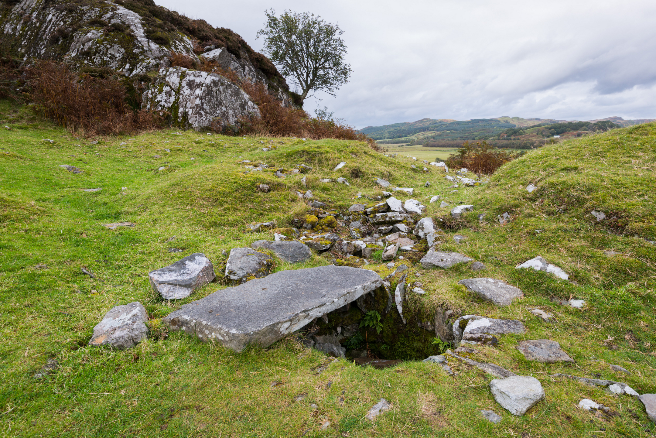 Broken stone slabs and a small stone-lined pit on grassy ground near rocky slopes at Dunadd Hill.
