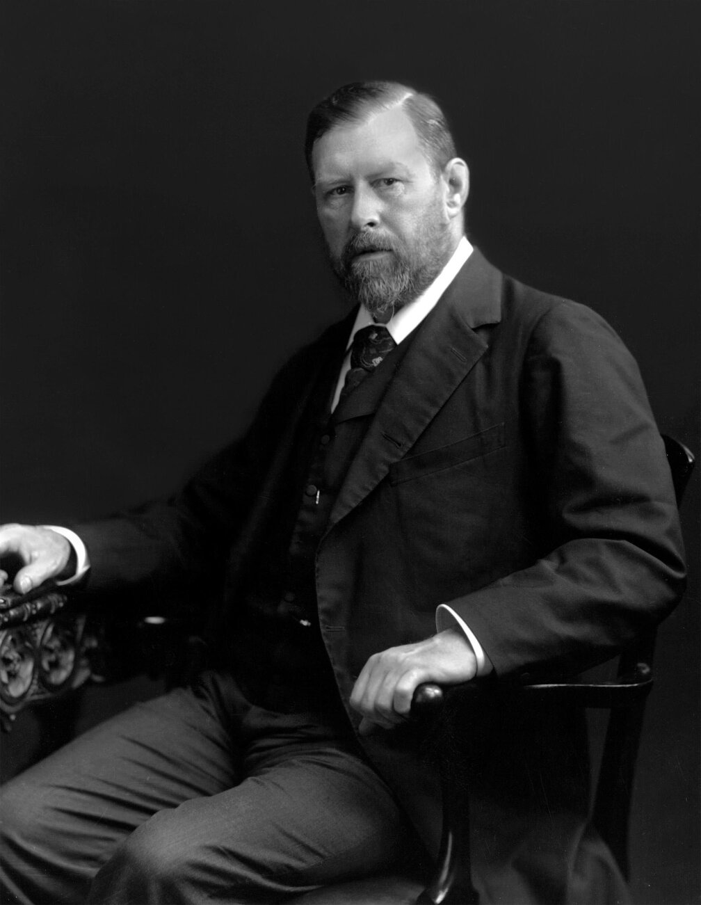 Black and white image of a seated man wearing a suit and tie. He has one hand on the arm of the chair and is looking at the camera.