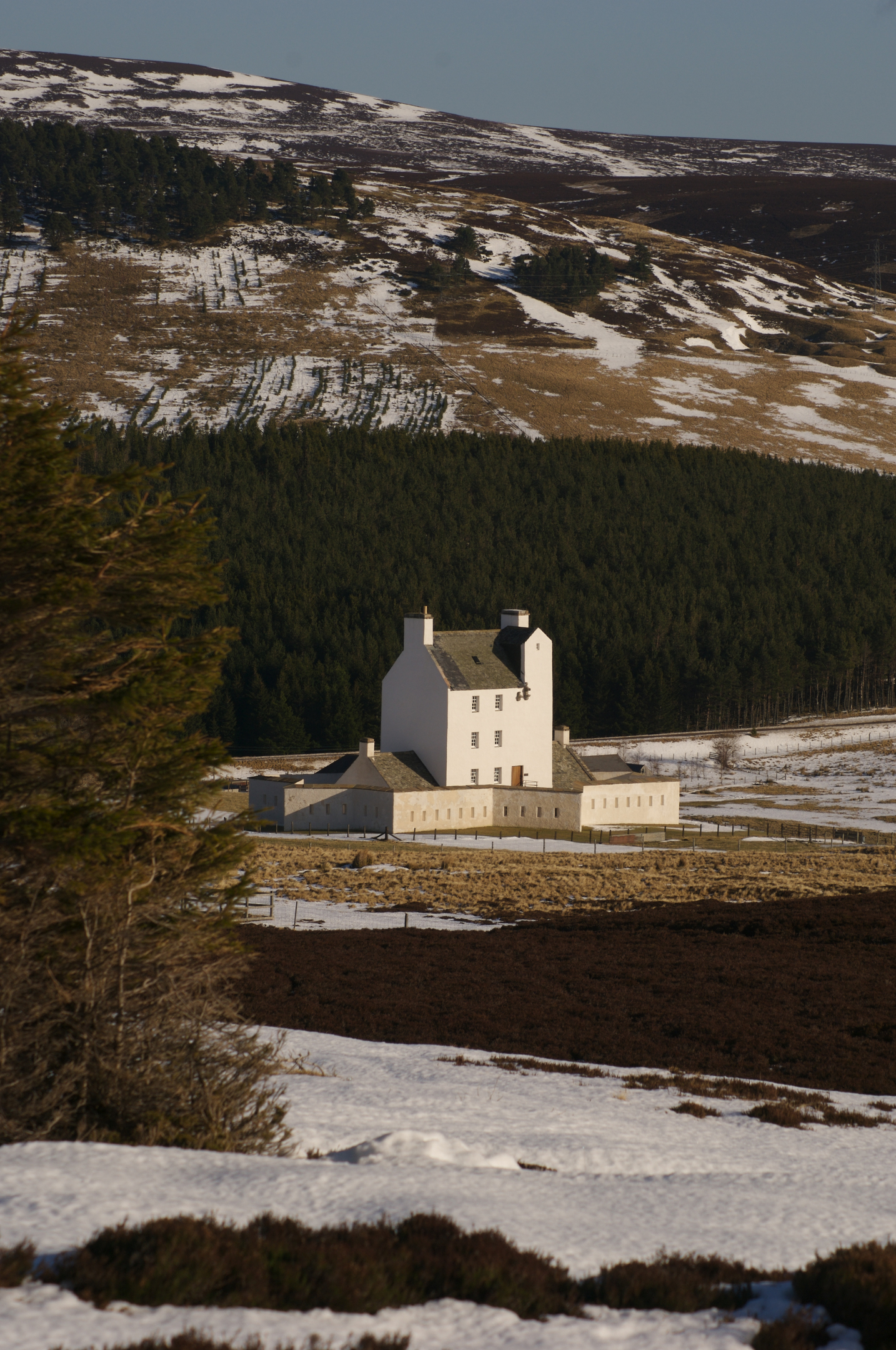 Corgarff Castle in a snowy valley, surrounded by forest and hills, with bright afternoon sunlight illuminating the building.