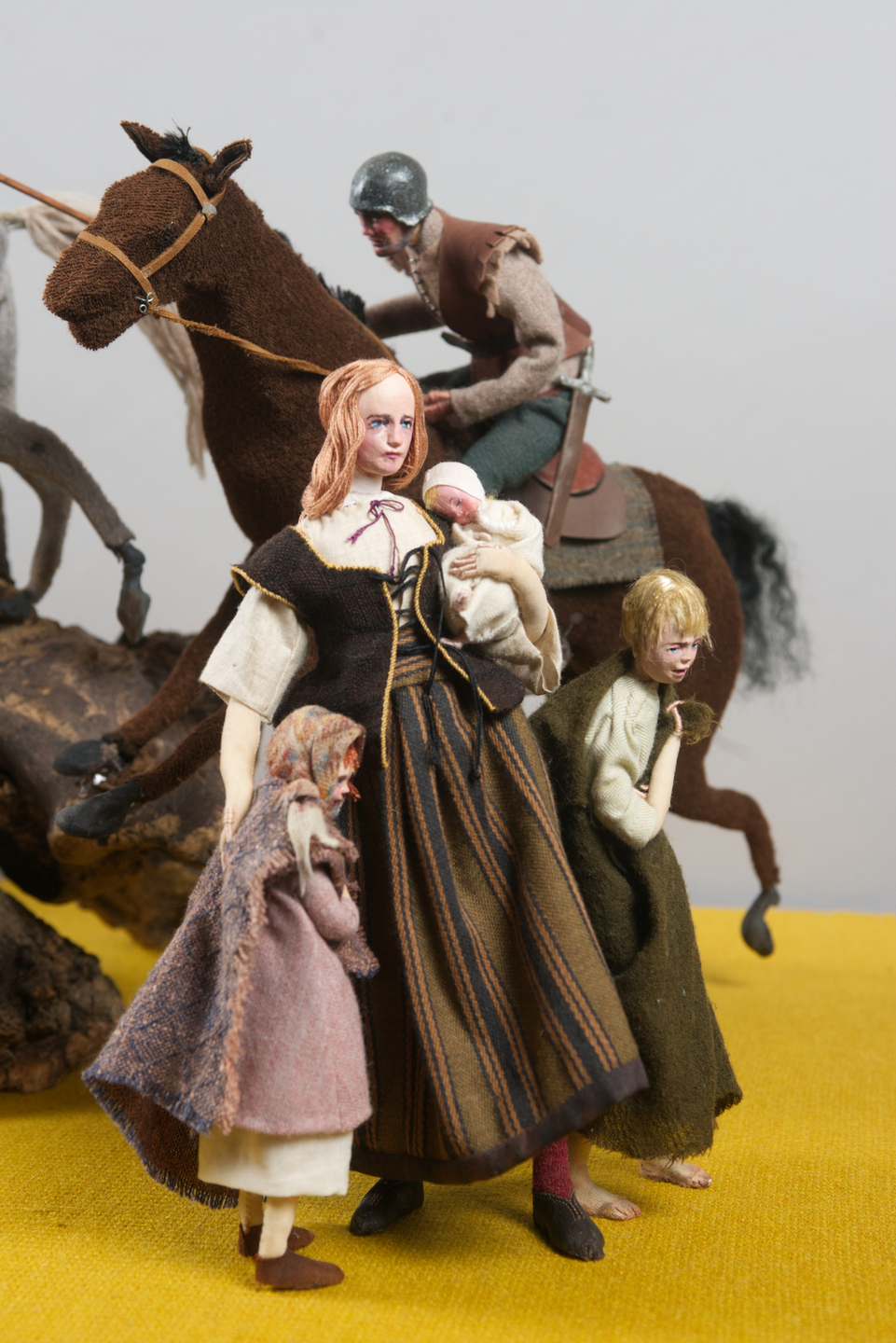 A group of four figures—an adult woman holding an infant, a young girl hugging her side, and an older woman leaning forward—stand in the foreground. Behind them, a mounted rider on a brown horse charges past, creating a sense of urgency or danger.