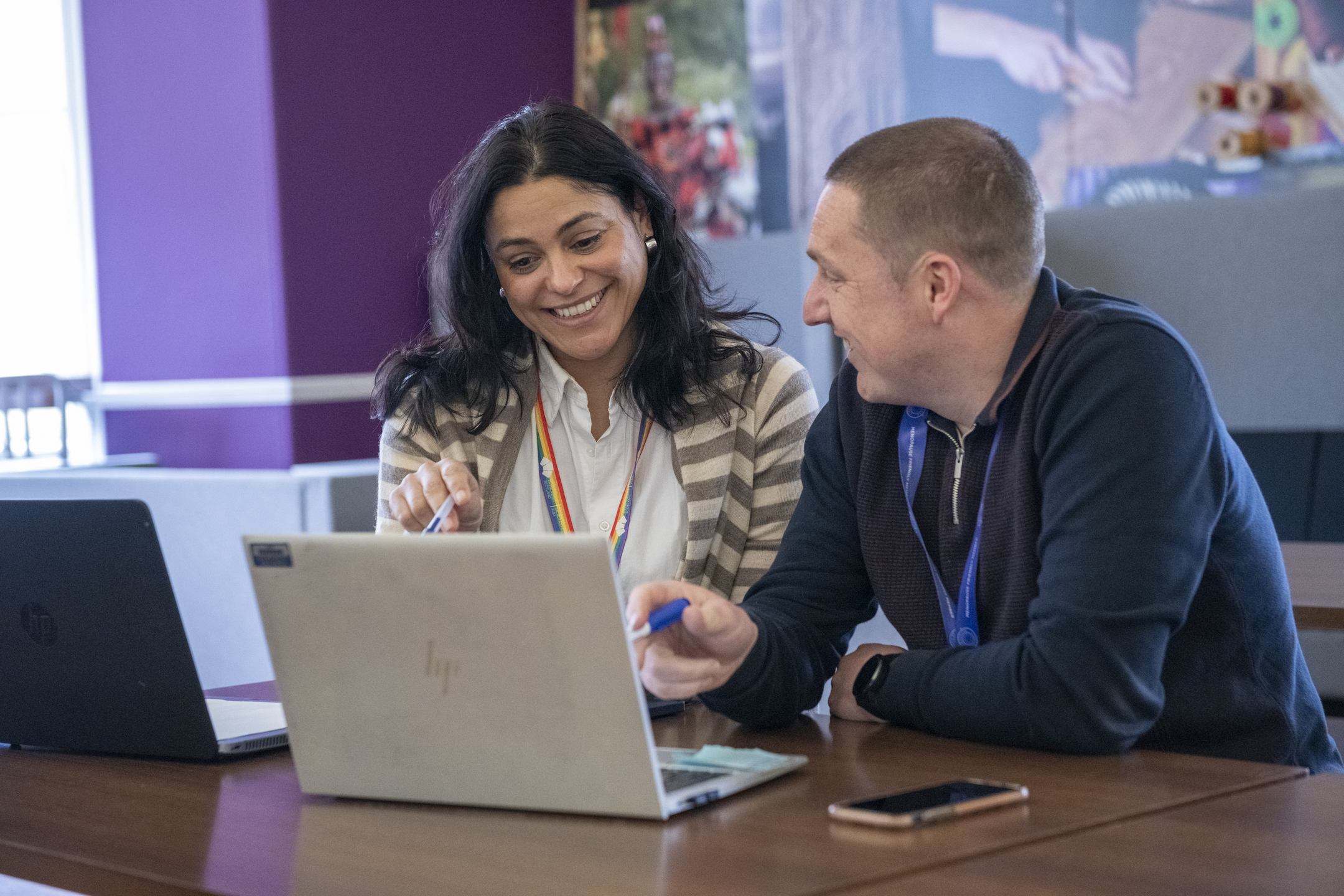 A man and a woman, both wearing Historic Environment Scotland lanyards, are sat looking at a laptop and smiling.
