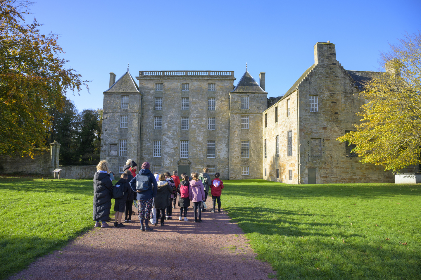 A group of young children, with a few adults, look up at a large stately home, with grey and brown stone. They stand on a path leading up to the building, with grass either side. There are two trees either side of the house. 