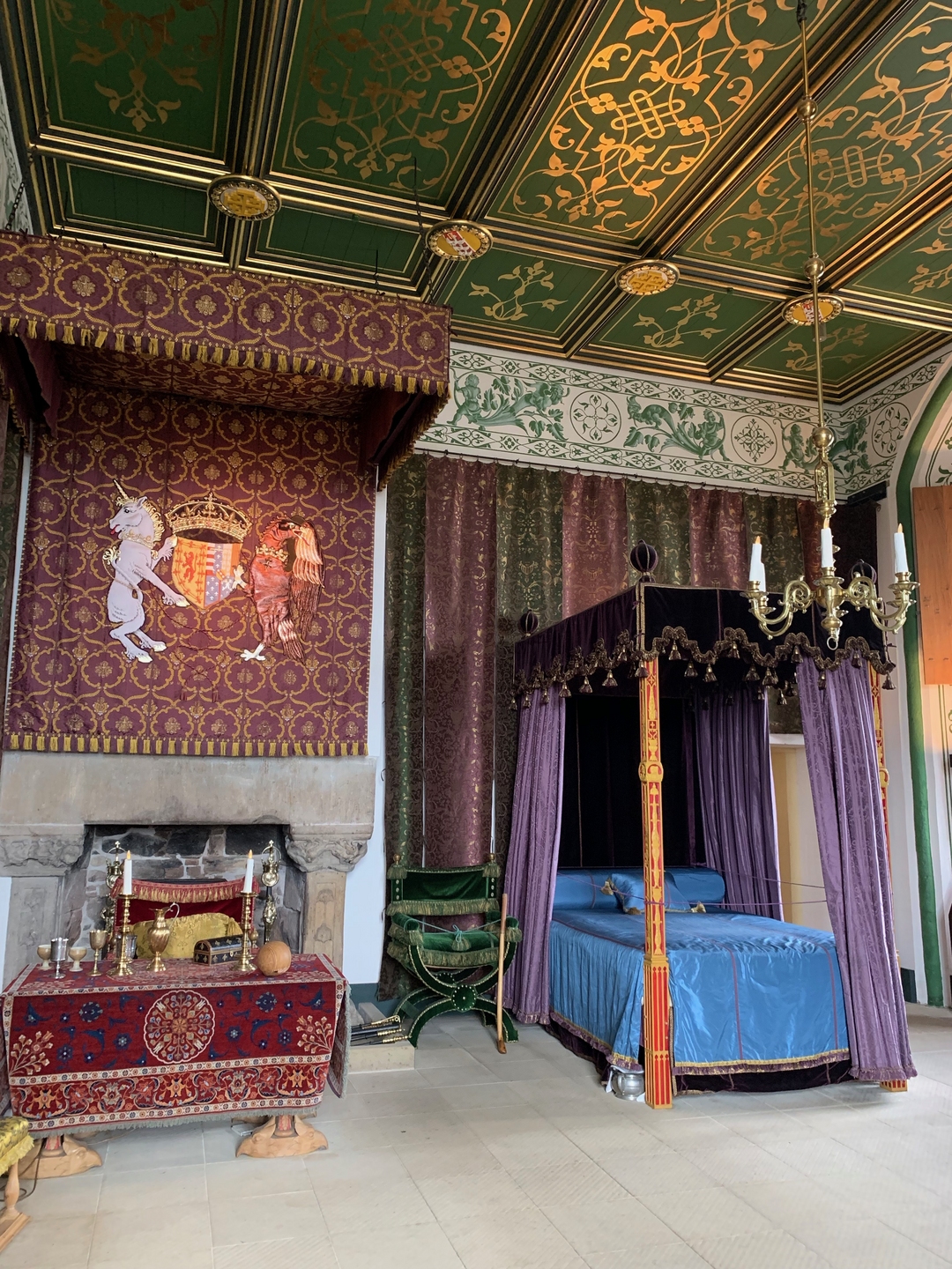 A furnished bedchamber, with hanging red embroidery, painted green and gold ceiling. There is a bed to the right with purple curtains, a hanging light and a table on the left with formal candles. 
