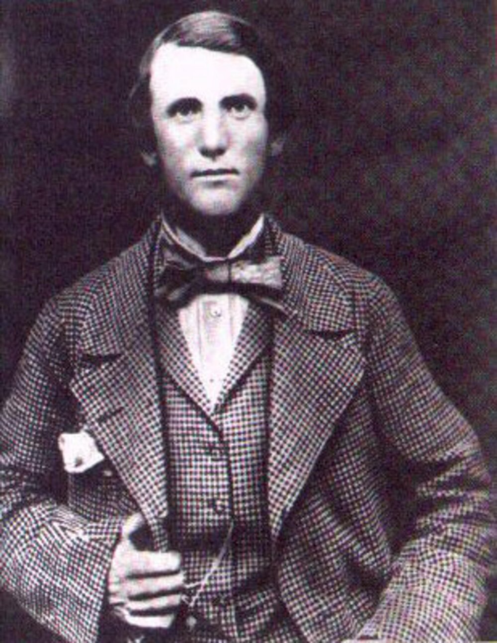 Black and white portrait photo of a person wearing a tweed suit and waistcoat. They also have a large dark bow tie and are looking at the camera with a relaxed expression, their right hand holding the lapel of their jacket.