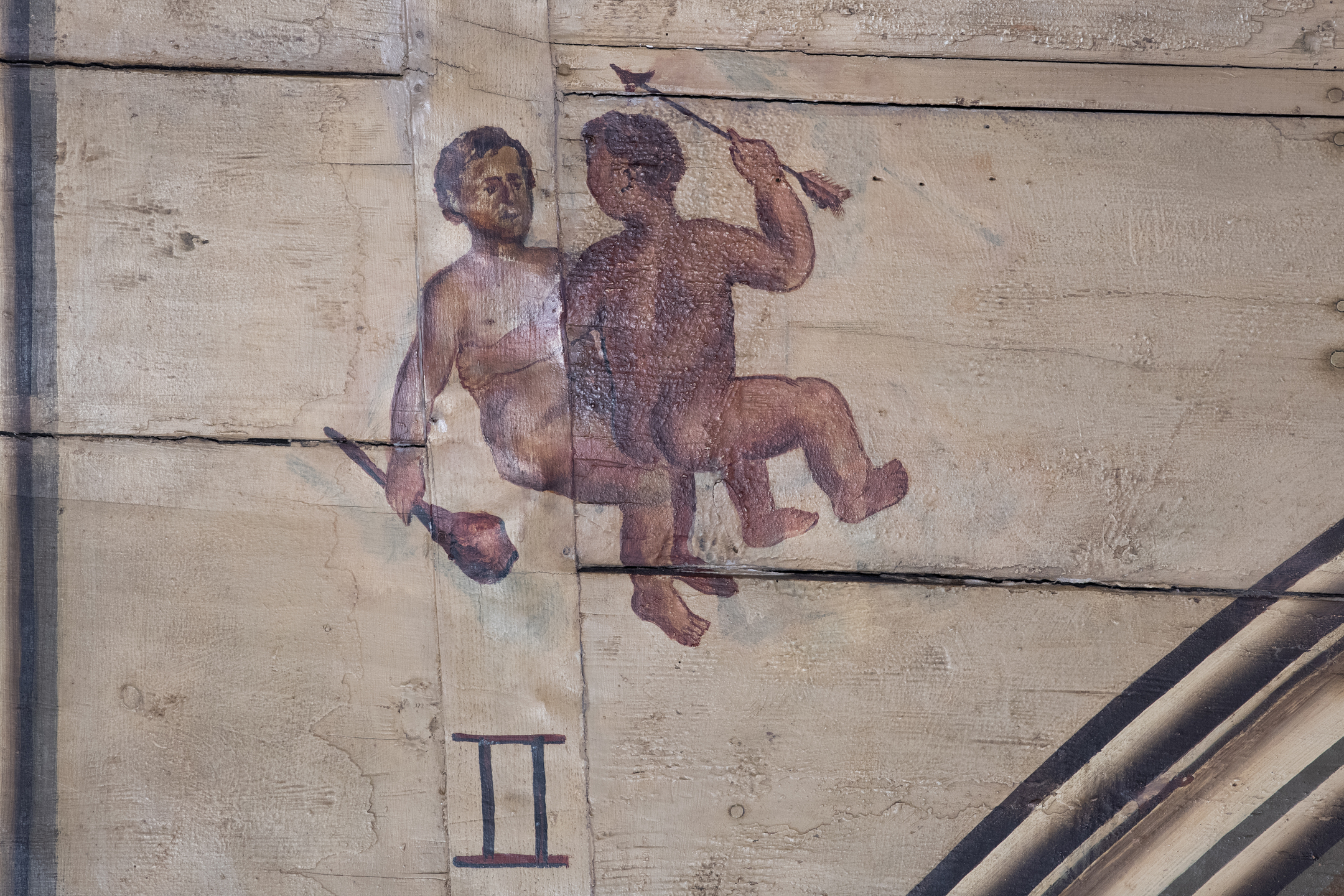 A painted depiction on aged wooden ceiling panels showing two small, childlike figures seated back‑to‑back, each holding a small staff or wand, with the Roman numeral II beneath them, representing the zodiac sign Gemini